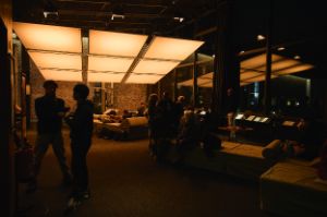 Eine großflächige Lichtinstallation an der Decke. Darunter Menschen auf Sofas oder im Raum verteilt.