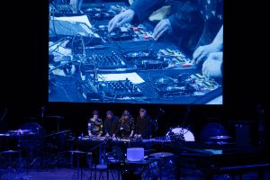 Das Ensemble arbeitet mit elektronischen Musikgeräten. Ihr Spiel wird zusätzlich über ihnen als Projektion gezeigt.