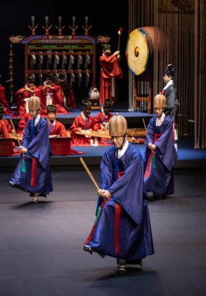 Männer in rituellen blauen Gewändern tanzen mit Holzschwertern in den Händen, hinter ihnen befindet sich ein Orchester in rituellen roten Gewändern.