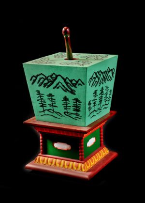 A green cube-like object painted with a landscape of mountains and trees is on a plinth.