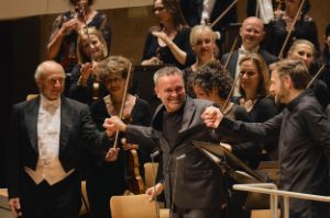Three people hold hands, smiling, orchestra musicians standing behind them.