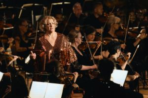 Alice Coote stands between the musicians of the orchestra in an orange sequined dress.