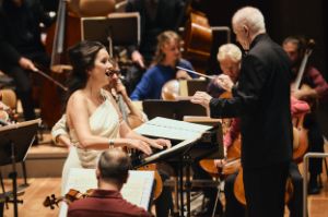 Anna Prohaska sings in a white dress in front of the Ensemble Modern Orchestra, Sir George Benjamin conducts next to her.