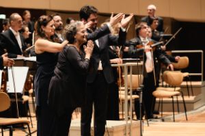 Three people stand in grateful hand positions in front of the standing orchestra.