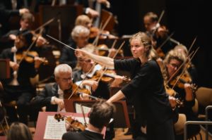 Mirga Gražinytė-Tyla conducts the orchestra with outstretched arms and baton in hand.