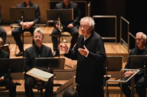 Philippe Herreweghe stands in front of the Collegium Vocale Gent, hands raised at chest level.