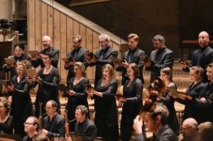 The choir sings and stands in two rows and elevated behind the orchestra.