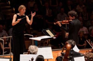 Joana Mallwitz holds the baton, facing the orchestra and Augustin Hadelich on the violin.