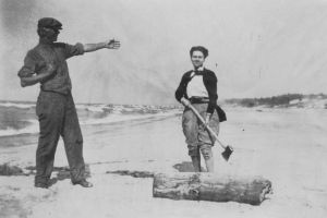 Ein Mann deutet auf eine Frau, die am Strand mit Beil über einem Baumstamm steht