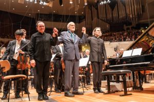 Drei Männer im Anzug stehen vor dem Orchester auf der Bühne und halten sich an den Händen.