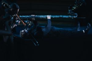Ein Mann mit einer Kontrabassklarinette liegt im Dunkeln auf der Bühne, aus dem Instrument spritzt etwas Glitzerndes hervor.
