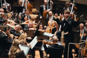 Ein Mann dirigiert ein Orchester und hebt gerade seine Hände hoch.