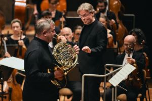 Ein Mann spielt auf dem Horn, während der Mann hinter ihm am Dirigentenpult steht.