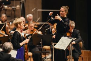 Eine Frau mit geschlossenen Augen hält eine Geige in der Hand, hinter ihr steht eine Frau und dirigiert das Orchester.