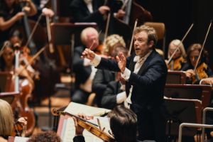 Ein Mann mit einem Taktstock in der gehobenen Hand steht vor den Streichern des Orchesters.