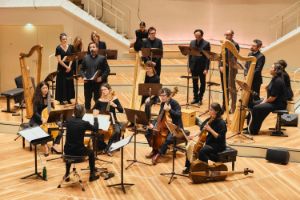 Auf der Bühne musizieren circa 15 Personen miteinander: Sänger*innen, Musiker*innen mit Streichinstrumenten und Harfen sowie ein Dirigent.