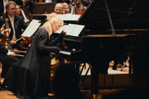 Eine hellblonde Frau in schwarzer langer Kleidung sitzt am Klavier, die rechte Hand auf den Tasten, die linke Hand vor ihrem Gesicht.