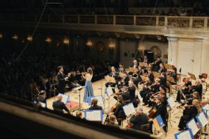 Blick von der Seite ins Publikum auf die Bühne, auf der Orchestermusiker, eine Violin-Solistin und ein Dirigent musizieren.