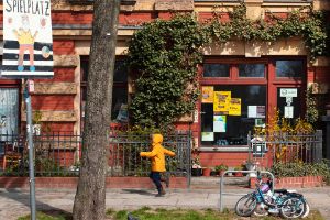 Eingang eines Ladens im Erdgeschoss eines Altbaus, davor ein Kind und das Schild „Spielplatz“.