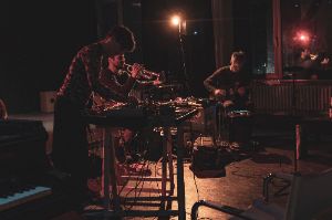 Three musicians with keyboard, trumpet and guitar play in a small dark room.