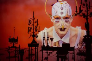 On a red-lit stage are chandeliers and a table, and performer Steven Cohen in a fancy costume is projected onto a large screen. 
