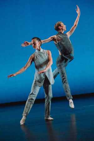 Two dancers in front of a light blue background. One person jumps into the air with their arms open, the other holds their arms towards an outstretched leg.