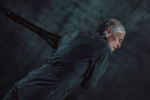 Lucinda Childs pulls on a rope. The choreographer wears a dark grey jumpsuit and has grey hair.