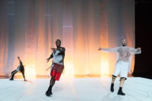 Three dancers in loose sportswear on a stage. The person in the background is in a supporting position.