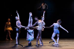 A group of dancers practising on a ballet barre. One person is standing on their head.