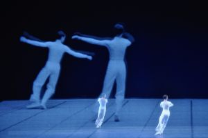 Zwei Tänzer:innen auf einer leeren Bühne. Im Hintergrund eine Projektion der zwei Tänzer:innen.