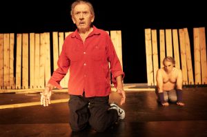 André Jung kneels in front of a fence made of vertically positioned wooden beams. He is wearing a shirt and gazing into the distance.
