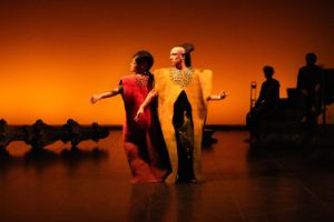 Two performers in long felt dresses. Behind them, silhouettes of musicians.