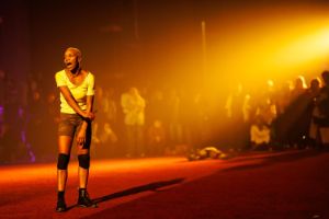 A person of Colour stands in a spotlight in the atrium, surrounded by an audience.