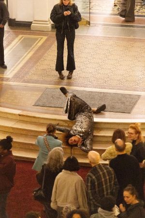 Eine weiblich gelesene Person in einem silbernen Kleid, liegt auf den Treppenstufen des Lichthofs im Gropius Bau und performt.