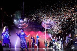 large disco balls and confetti above dancing performers dressed in striking costumes