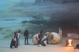 Actors in a dystopian stage landscape look into an oversized egg.