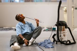 A person of Color with funky glasses and barbecue tongs, next to a vacuum cleaner