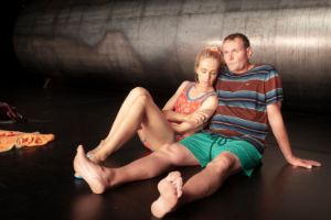 Two actors with sad faces sit on the stage leaning against each other.