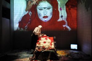 Live projection on stage: a make-up artist paints a person's face with mask-like and ornamental elements.