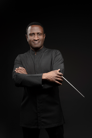 A man wearing a black suit and holding a baton looks kindly into the camera.