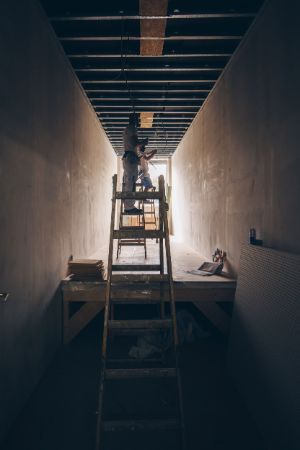 A corridor with an open ceiling and three ladders behind each other. There is one person in work clothes on each of the ladders at the back.