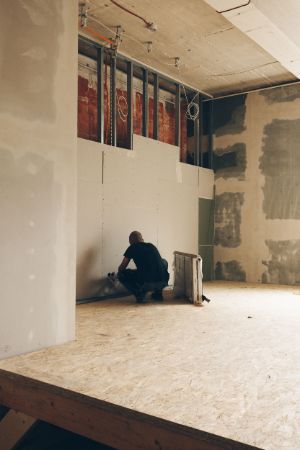 A man squats in a gutted room in front of a wall that is covered in the lower half. Steel beams are visible in the upper half.