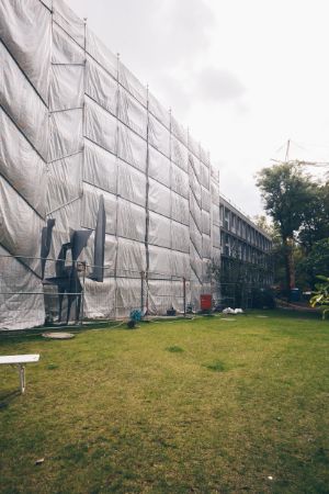 View of the outer wall of a building covered with light-coloured foil with construction fences in front of it - a green meadow can be seen in the foreground.