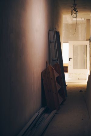 View into a darkened corridor with various objects leaning against the unpainted wall. A door that has been lifted off its hinges leans against the brightly lit end of the corridor.