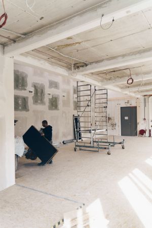 In a gutted hall with scaffolding on wheels is a man carrying a large black plate.