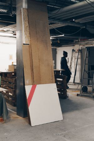 Ein kellerartiger Raum mit einer Säule, an der Spanplatten angelehnt sind. Neben verschiedenen Leitern und Objekten ist im Hintergrund ein Mann erkennbar.