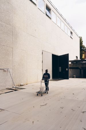 In front of the slab-clad outer wall of a building with an open door, a man pushes a luggage cart on which a bucket is placed.