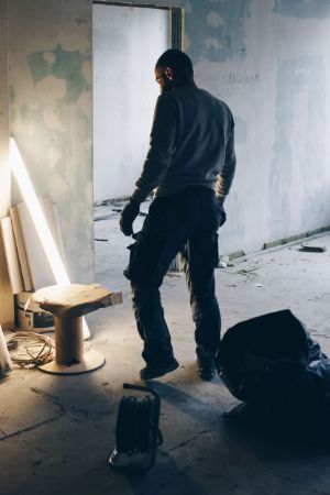 In a gutted room with cable reels and a rubbish bag, among other things, a fluorescent tube leans against the wall, somewhat illuminating the face of a man in work clothes.