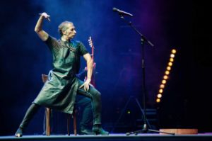 Ein Tänzer in einem dunkelgrünen Kleid mit erhobener rechter Hand steht vor einem Mikrofon auf der Bühne. Größtenteils verdeckt dahinter ein Gitarrenspieler.
