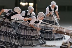A group of four women in traditional costumes knot a rope to a wooden beam.
